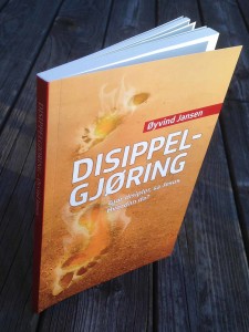 En bok som står oppreist på gamle treplanker. Bokens forside er av dype fotspor i sand, det ryker og gløder fra fotsporene. Tittelen på boken er "Disippelgjøring" av Øyvind jansen. Undertittelen er: "Gjør disipler, sa Jesus. Hvordan da?"